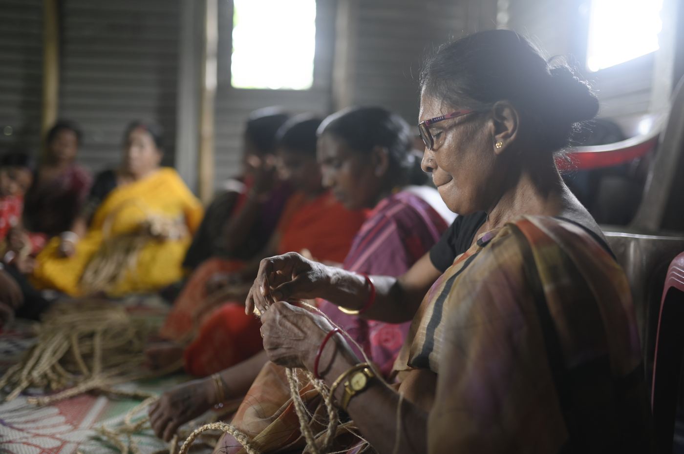 A senior artisan in spectacles twists raw jute fibre between her fingers, other women working behind her